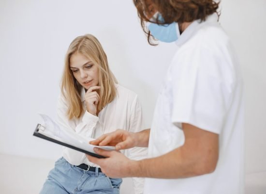 A doctor in a face mask discusses diagnosis with a patient in a clinic setting.